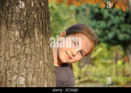 Blanc caucasien 8 ans enfant, fille, heureux dans le parc, souriant et se cachant derrière un grand arbre Banque D'Images