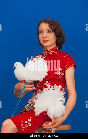 Une belle femme vêtue d'une robe rouge est assise avec une paire d'amoureux de pigeon blanc dans ses mains sur un fond bleu. Photo de haute qualité Banque D'Images