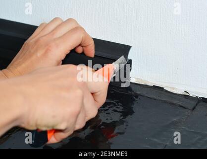 Un homme de main installe une plinthe en plastique noir sur un mur blanc en le coupant à l'aide d'un couteau universel. Banque D'Images