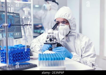 Technicien de laboratoire en équipement de protection individuelle examinant des échantillons de virus au microscope en laboratoire. Scientifique en costume de protection assis sur le lieu de travail utilisant la technologie médicale moderne pendant l'épidémie mondiale. Banque D'Images