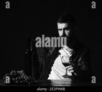 Un homme avec une barbe tient un verre de vin sur fond marron foncé. Le sommelier goûte des boissons chères. Concept de dégustation de vins et de dégustation. Le Degustator au visage grincheux se trouve à côté d'une bouteille de vin et de raisins foncés. Banque D'Images