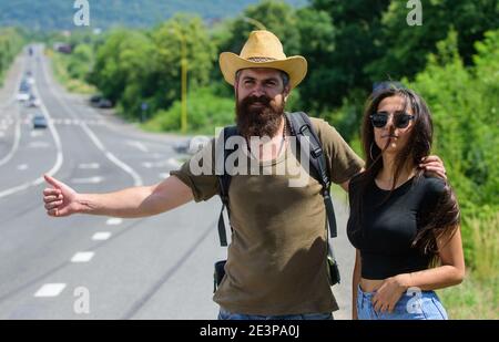 Couple voyageurs homme et fille hitchranking à bord de route nature arrière-plan. Les voyageurs essaient d'arrêter leur voiture. La randonnée est l'un des moyens les moins chers de voyager. Couple de randonneurs voyageant l'été ensoleillé jour. Banque D'Images