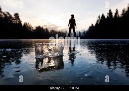 Un cube de glace clair et son reflet sur un lac gelé plat, au lever du soleil, éclairé par le soleil depuis l'arrière-plan, avec une femme patinant sur glace en arrière-plan, la Slovénie Banque D'Images