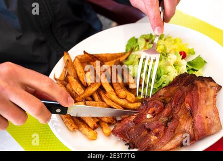 Un homme qui travaille dur pour un déjeuner nutritif. Poitrine de porc grillée, frites et salade verte. Banque D'Images