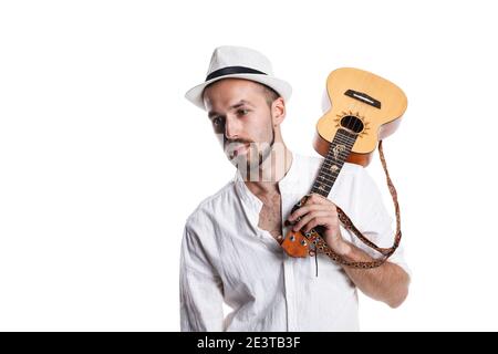 Jeune homme caucasien attrayant avec ukulele isolé sur fond blanc .Homme en blanc chapeau d'été et chemise avec ukulele pieds nus en studio blanc pho Banque D'Images