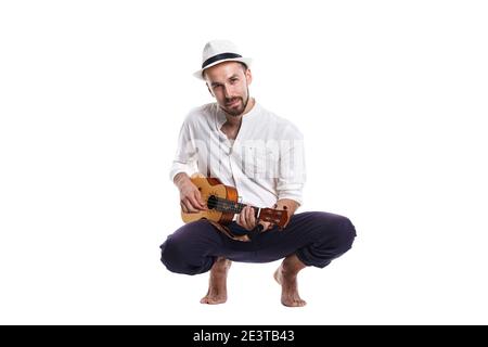 Jeune homme caucasien attrayant avec ukulele isolé sur fond blanc .Homme en blanc chapeau d'été et chemise avec ukulele pieds nus en studio blanc pho Banque D'Images