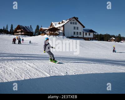 Un gars dans un casque descend la montagne sur un snowboard avec la toile de fond des bâtiments de la station de ski lors d'une journée d'hiver ensoleillée. Banque D'Images