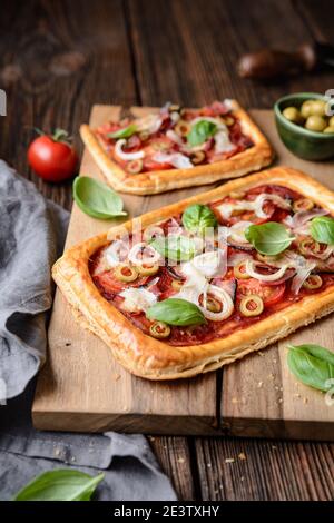 Pizza feuilletée maison avec tranches de bacon, tomates, olives vertes, fromage et oignons, recouvertes de feuilles de basilic sur fond rustique en bois Banque D'Images
