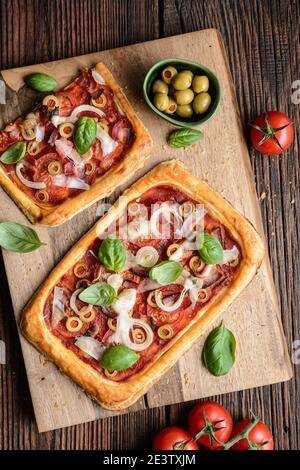 Pizza feuilletée maison avec tranches de bacon, tomates, olives vertes, fromage et oignons, recouvertes de feuilles de basilic sur fond rustique en bois Banque D'Images