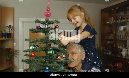 Petite fille d'enfant avec grand-parent senior décorant l'arbre de Noël artificiel à la maison. Intérieur de salon de style ancien. Grand-père âgé avec un enfant célébrant les vacances en famille du nouvel an Banque D'Images