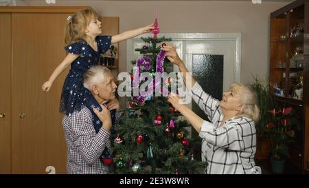 Petite fille d'enfant avec grands-parents de famille décorant l'arbre de Noël artificiel à la maison. Intérieur de salon de style ancien. Grand-mère, grand-père et enfant âgés célébrant les fêtes du nouvel an Banque D'Images