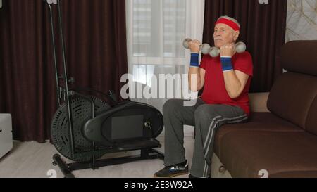 Vieux modèle de grand-père senior aux cheveux gris faisant des exercices de levage de poids haltères dans la chambre à la maison. Style rétro Caucasien mature homme âgé se réchauffant tout en étant assis sur le canapé. Un mode de vie sportif sain Banque D'Images