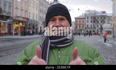 Portrait de l'homme expérimenté élégant touriste souriant, montrant le pouce vers le haut, regardant l'appareil photo dans le centre-ville d'hiver de Lviv, Ukraine. Photographie, voyage, vacances. Les retraités actifs vient après leur retraite Banque D'Images