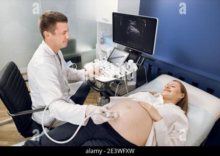 Souriant médecin obstétricien, examinant le ventre de la femme enceinte heureuse par balayage ultrasonique. Gai homme médecin faisant l'échographie procédure pour son Banque D'Images