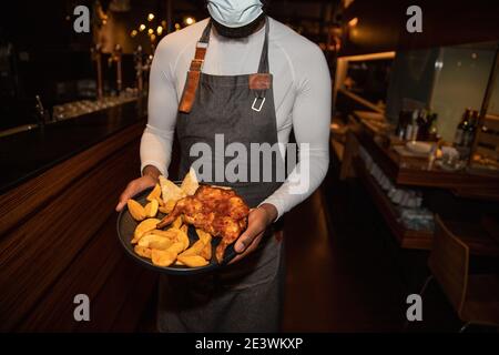 Un serveur africain avec une assiette de poulet et de pommes de terre porte un masque de coronavirus pour se protéger contre la pandémie de coronavirus Banque D'Images