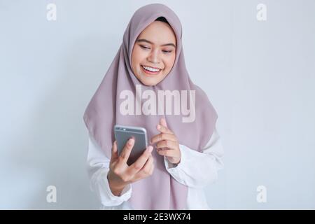 La jeune femme asiatique de l'Islam portant un foulard est souriante et heureuse dans ce qu'elle voit sur le smartphone. Femme indonésienne sur fond gris. Banque D'Images