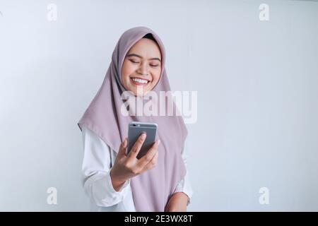 La jeune femme asiatique de l'Islam portant un foulard est souriante et heureuse dans ce qu'elle voit sur le smartphone. Femme indonésienne sur fond gris. Banque D'Images