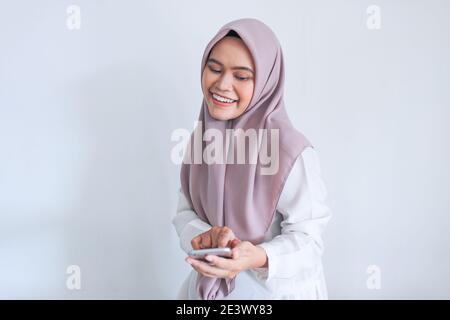 La jeune femme asiatique de l'Islam portant un foulard est souriante et heureuse dans ce qu'elle voit sur le smartphone. Femme indonésienne sur fond gris. Banque D'Images