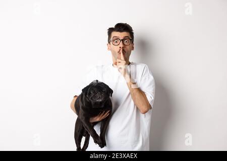 Image d'un jeune homme drôle tenant un mignon chien de pug noir et se précipitant à l'appareil photo, montrant le signe de hush et regardant à l'appareil photo, en gardant un secret, se tenant dessus Banque D'Images