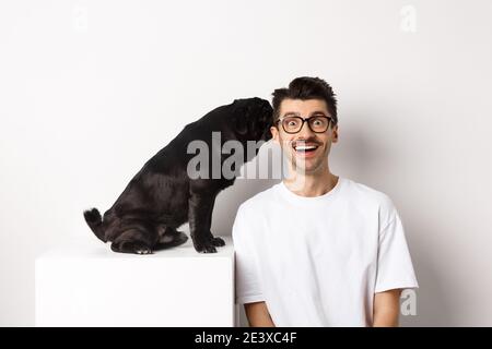 Image d'un joli chien de boug noir qui murmure à l'oreille du propriétaire, homme qui a l'air étonné et souriant, debout sur fond blanc Banque D'Images