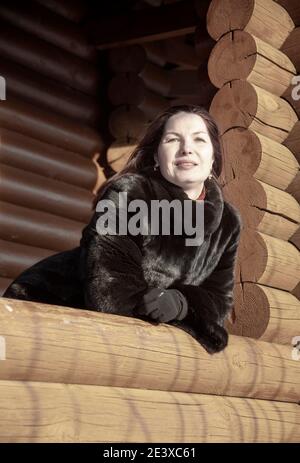 Une belle femme de 45 ans aux cheveux rouges pose sur la véranda d'une maison en rondins le jour d'hiver. Banque D'Images