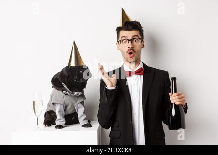 Drôle de maque noire dans le cône de fête mettant en vedette le maître de chien surpris, célébrant l'anniversaire, boire du champagne et porter des costumes, fond blanc Banque D'Images