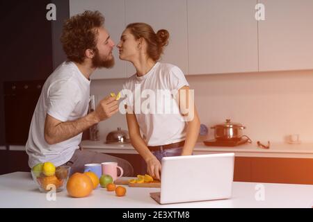 De beaux jeunes couples s'embrassant se nourrissant les uns les autres, se faisant plaisir dans une cuisine moderne et souriant tout en cuisinant à la maison. Jolie fille nourrit ou lui allaite Banque D'Images