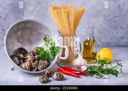 sur fond gris, dans une vieille passoire, des palourdes fraîches à la carapace, des spaghetti et divers ingrédients pour la préparation des pâtes. Toujours Banque D'Images