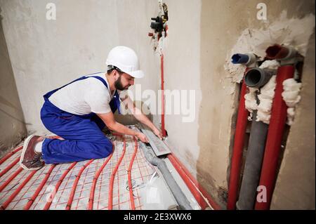 Vue grand angle sur un jeune travailleur debout sur ses genoux dans une combinaison bleue et un casque installant un système de chauffage au sol et un système de plomberie. Concept de rénovation de la maison et travaux de plomberie. Banque D'Images