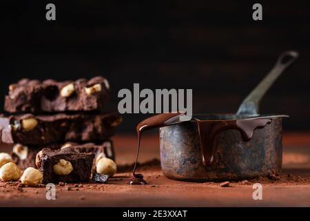 Au premier plan quelques morceaux de chocolat noir empilés avec des noisettes. À part la casserole en cuivre avec le chocolat fondu et le spoo goutte à goutte Banque D'Images