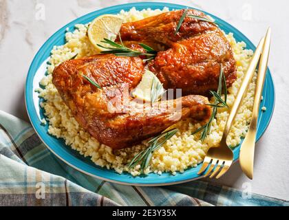 Des quartiers de cuisses de poulet servis sur le millet sur une assiette avec des couverts dorés, des quartiers de citron et des brins de romarin frais sur un fond de pierre en marbre blanc Banque D'Images