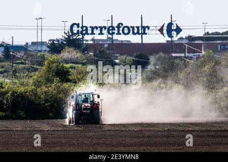 Agriculteur travaillant dans un champ avec son tracteur et Carrefour supermarché en arrière-plan Banque D'Images