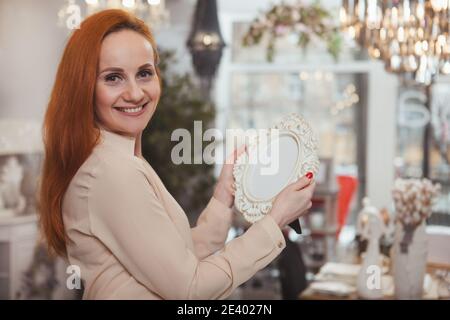 Bonne femme rouge aux cheveux souriant à l'appareil photo au-dessus de son tibia, tenant le cadre photo. Belle femme mature magasiner à la maison décoration magasin. Femme de ménage, moi Banque D'Images