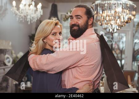 Un couple d'âge mûr souriant à l'appareil photo, tenant des sacs d'achats. Beau barbu homme mûr embrassant sa belle femme tout en faisant du shopping ensemble. Réf Banque D'Images