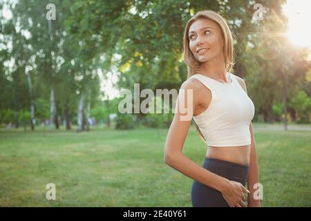 Magnifique femme heureuse et en bonne santé avec corps en forme souriant, marchant dans le parc au coucher du soleil, espace de copie. Femme sportive gaie dans des vêtements de sport appréciant le rel Banque D'Images