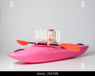 Photo studio d'un bébé assis dans un kayak rose avec une palette orange, sur fond blanc Banque D'Images