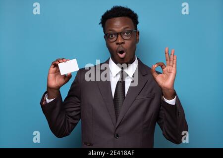 Jeune homme d'affaires africain sérieux tenant une carte de crédit montrant le geste ok. Studio tourné sur un mur bleu. Banque D'Images