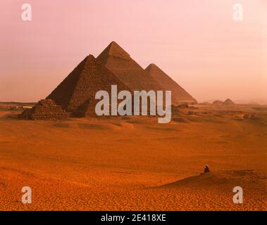Afrique du Nord Égypte le Caire pyramides de Gizeh au coucher du soleil heure dorée lever du soleil dans le paysage désertique avec l'homme assis dans le désert Banque D'Images