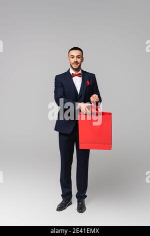 vue sur toute la longueur de l'homme hispanique heureux en costume élégant mettre le cadeau dans le sac sur fond gris Banque D'Images