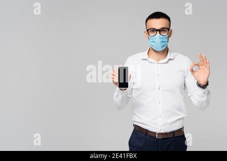 manager hispanique dans le masque médical montrant un geste correct et un smartphone avec écran vierge isolé sur gris Banque D'Images