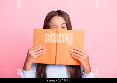 Portrait en gros plan d'une jolie petite fille aux cheveux bruns qui se cache derrière livre en clin d'œil isolé sur fond rose pastel Banque D'Images