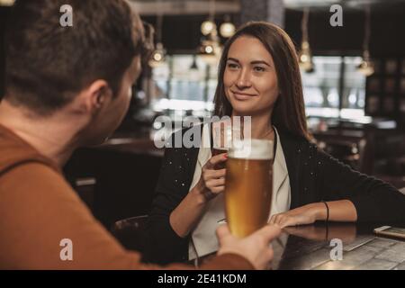 Belle femme heureuse souriant gaiement, parlant à son petit ami avec un verre de bière au pub local. Une jeune femme charmante boit un délicieux artisanat Banque D'Images