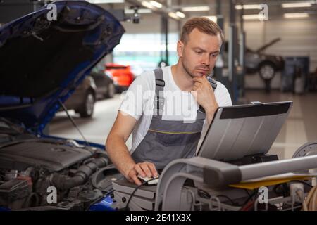 Mécanicien de voiture mûr regardant concentré, en utilisant l'ordinateur portable tout en réparant une voiture au garage. Technicien automobile expérimenté utilisant l'ordinateur dans son atelier Banque D'Images