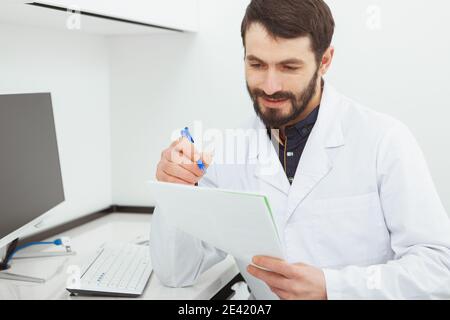 Gai homme mature médecin souriant en lisant des documents médicaux à son bureau. Praticien qui fait de la paperasserie à l'hôpital Banque D'Images