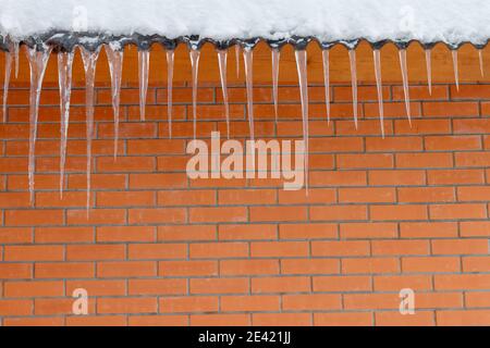 Une rangée de glaçons rassemblés sur un toit en face d'un mur de briques rouges. Morceaux de glace de différentes tailles accrochés sur un toit recouvert de neige, une surface en briques rouges sur le sol arrière. Photo de haute qualité Banque D'Images