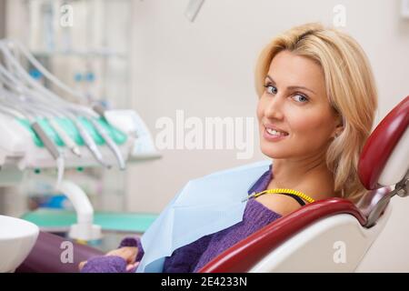 Décontractée belle femme souriant sur son épaule à la caméra, assise dans une chaise dentaire, en attente d'un rendez-vous médical avec son dentiste. Joyeux pati Banque D'Images