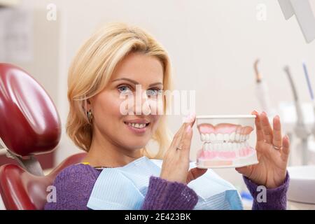 Belle femme mûre heureuse avec des dents blanches saines souriant joyeusement, assis dans une chaise dentaire à la clinique. Femme attrayante tenant le moule dentaire, waiti Banque D'Images