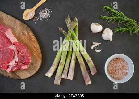 Asperges crues, romarin, steak cru, ail et sel de mer sur béton, fond de pierre. Concept de Flat Lay, vue de dessus. Alimentation saine et biologique. Banque D'Images