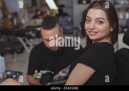 Gros plan d'une belle femme heureuse souriant à l'appareil photo tout en obtenant le tatouage de manchon de bras fait par le tatouage professionnel. Client féminin gai étant t Banque D'Images