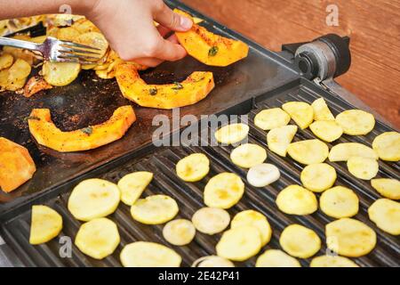 Morceaux de courge de noyer cendré d'orange vif grillés sur le gril électrique, les mains de la femme déplaçant des légumes, des chips de pomme de terre floues également cuites au premier plan Banque D'Images